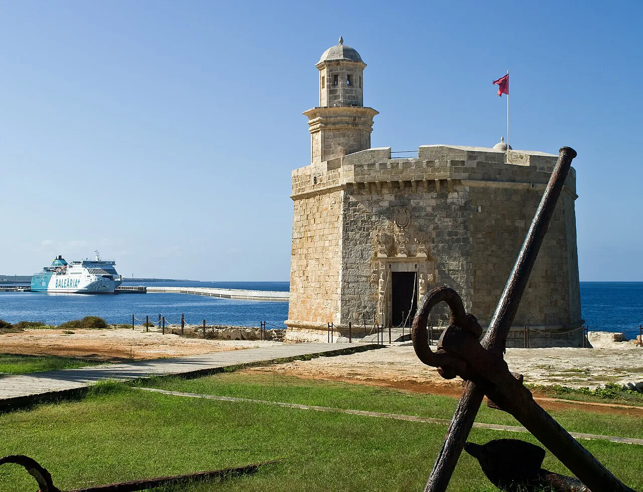 Castell de Sant Nicolau in Ciutadella, with an anchor on the lawn and a ferry offshore.